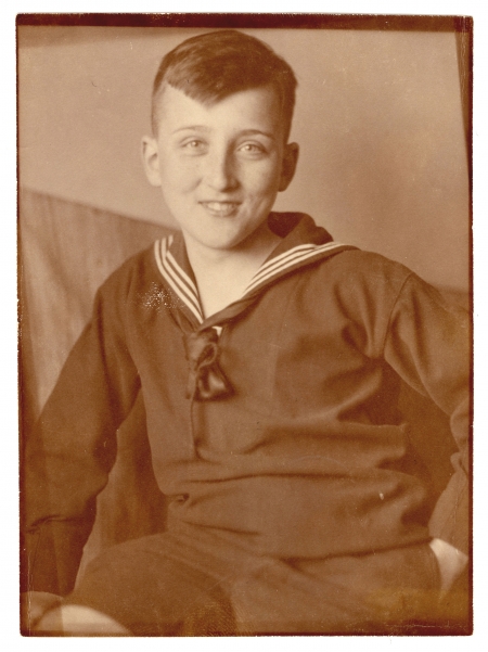 Sepia-toned photograph of an adolescent boy sitting and smiling at the camera. He wears a dark top with a sailor collar.