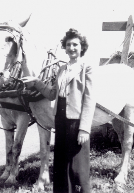 Photo en noir et blanc d'une femme aux cheveux bruns foncés, se tenant à l'extérieur auprès de deux chevaux blanc attachés à une voiture ou un charriot. Elle tient les rênes des cheveux d'une main.