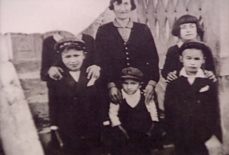 Black-and-white photograph of a woman with five children standing together, the three in the back row putting their hands on their family members’ shoulders in front. The photograph is torn in the top-left corner so that the face of the person standing in the top left is not visible.