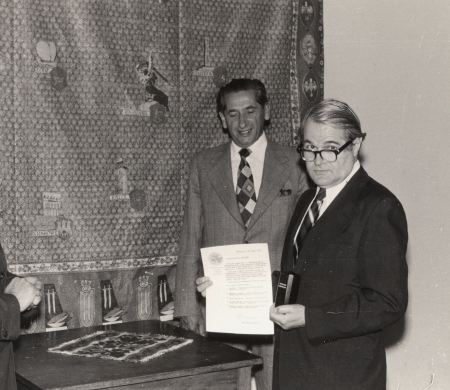 Black-and-white photograph depicting two men in suits standing around a table indoors. The man on the right is holding a document and a small urn.