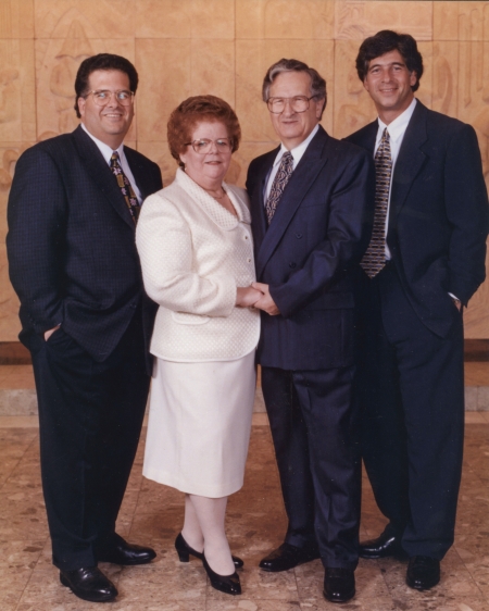 Colour studio photograph of a group of four adults standing together and smiling at the camera. The couple in the middle is elderly, and the woman wears a white jacket and skirt. The three men wear suits.