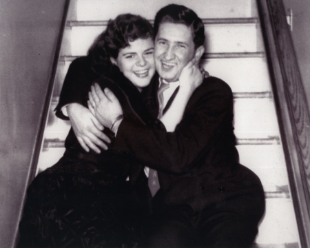 Black-and-white photograph of a young couple smiling and sitting in each other’s arms on a stairwell.