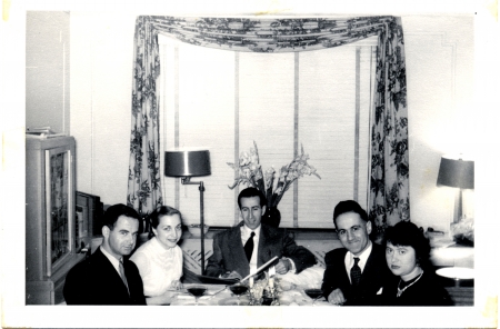 Black-and-white photograph of a group of 5 adults casually sitting at a dining table with glasses of wine, smiling at the camera. The men wear suits.