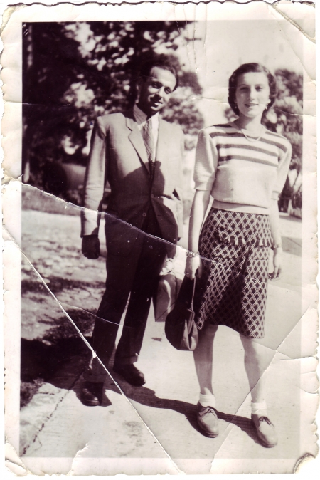 Black-and-white photograph of a man and woman standing together outdoors on the pavement, with trees in the background. The man wears a suit, and the woman wears a blouse and polka-dot skirt.