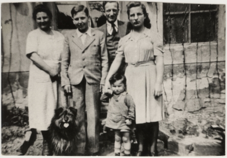 Black-and-white photograph of a man and woman standing outside the front of a house with two adolescent children, a young toddler child, and a dog.