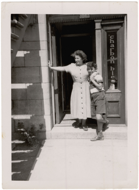 Black-and-white photograph of a woman and a boy standing at the doorstep of a ground floor apartment. The woman has her arm around the son and rests her other arm against the entrance to the apartment.