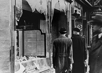 Black-and-white photograph of three men in hats and jackets, standing on a sidewalk and looking at two shattered storefront windows. Two men have their back to the camera.
