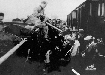 Black-and-white photograph of a man sitting on the edge of a train car, holding a cane, and looking onto a platform full of people.