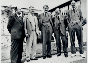 Black-and-white photograph of five men, wearing suits, standing outside in a row and smiling or laughing.