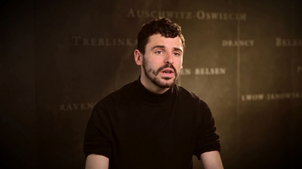 Capture d'écran de l'entrevue d'Antoine Burgard. Il est assis devant un mur gris sur lequel est inscrit les noms de divers camps de concentration. Son visage et ses épaules sont visibles à la caméra.
