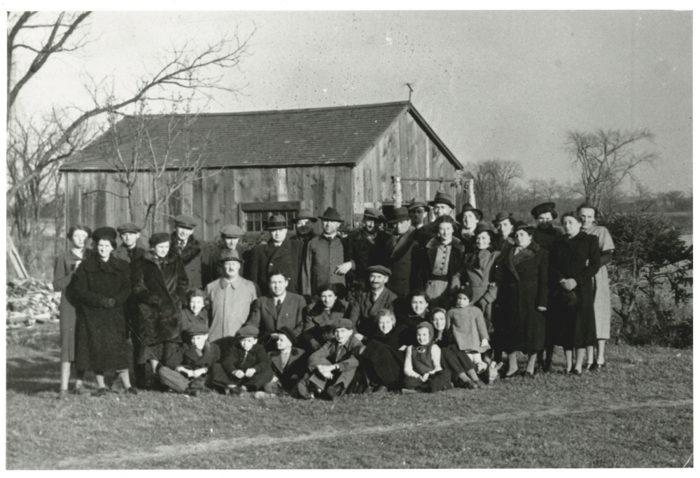 Photo en noir et blanc d'un grand groupe d'environ 35 hommes, femmes et enfants, habillés de manteaux d'hiver et de chapeau, rassemblés ensemble à l'extérieur sur une pelouse. Il y a une grange derrière le groupe en arrière-plan.