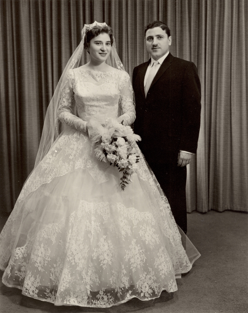 Black-and-white studio portrait photograph of a man and a woman on their wedding day. The woman wears a long white gown with a veil, holding a bouquet of flowers, and the man wears a formal suit and a moustache.