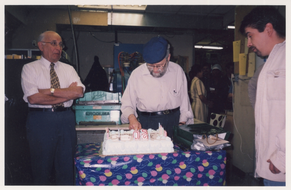 Colour photograph of an elderly man with a beard and cap, cutting into a large cake while two men stand on either side watching him.