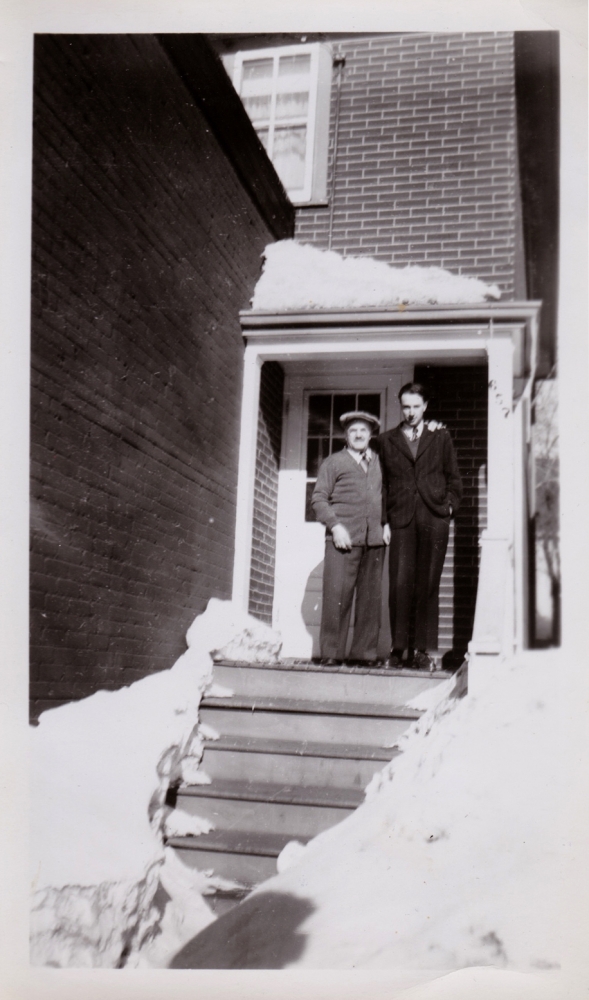 Black-and-white photograph of two men standing on the front porch of a brick house with snow in the foreground. Both men wear su