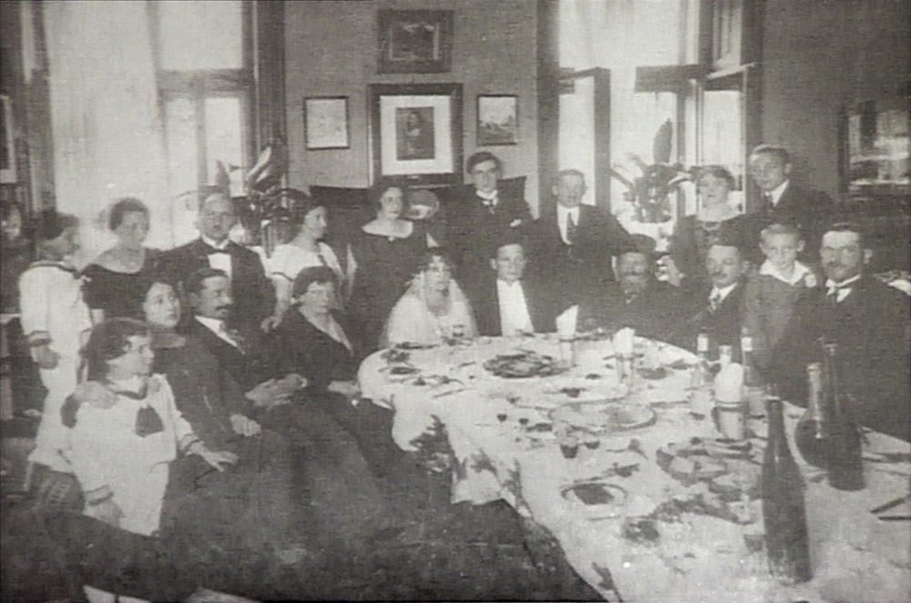 Photo en noir et blanc d'un groupe d'environ 20 personnes assises ensemble autour d'une grande table remplie de plats. Le groupe est habillé de manière formelle et un couple assis à la table semble célébré leur mariage.