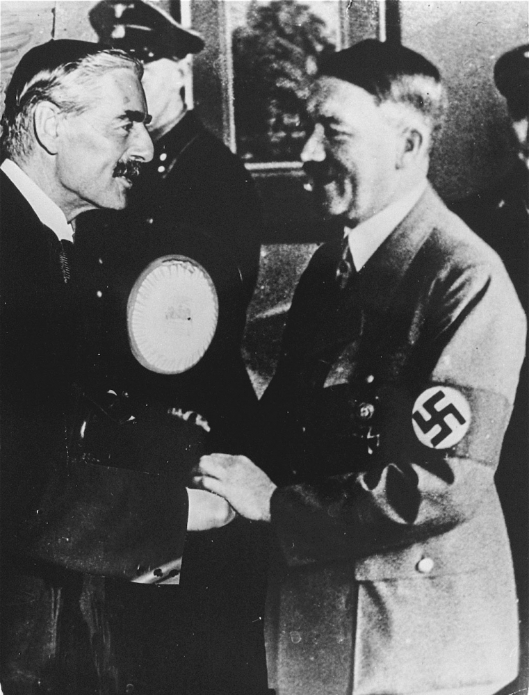 Black-and-white photograph of two men smiling and shaking hands. Both men have moustaches, and one man wears a lighter suit with a swastika badge on his arm.