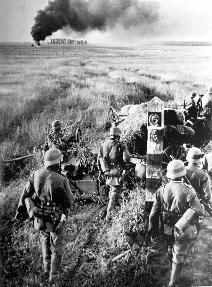 Photo en noir et blanc d'une troupe militaire d'environ une demi-douzaine d'hommes marchant dans un champ. Il semble y avoir un grand feu avec un grand nuage de fumée en arrière-plan.