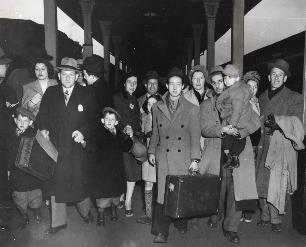 Black-and-white photograph a group of about 16 people walking towards the camera on a train platform. They are wearing jackets and some carrying suitcases. There are four young children in the group.