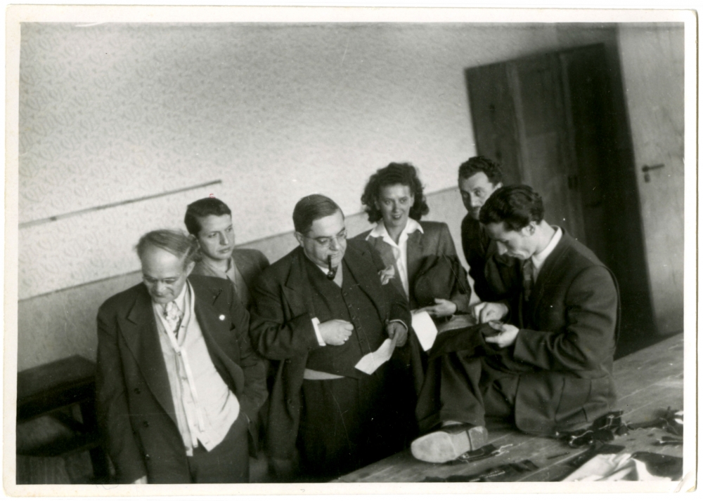 Photo en noir et blanc d'un groupe de six personnes, cinq hommes et une femme, se tenant debout dans une pièce. Un homme fume la pipe et observe un autre homme assis à une table qui cout un tissus à la main.