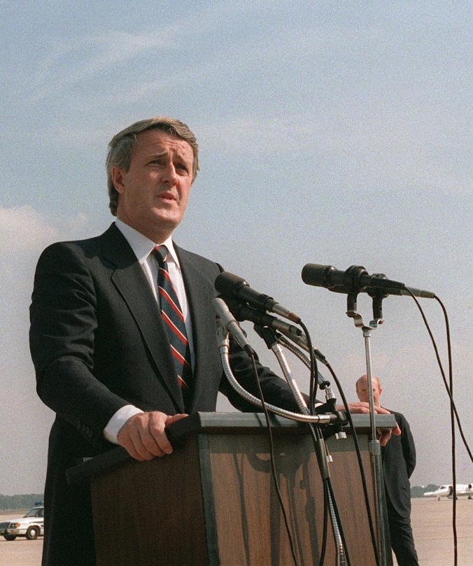 Colour photograph of a man in a suit, speaking outdoors at a podium hooked up with two microphones.