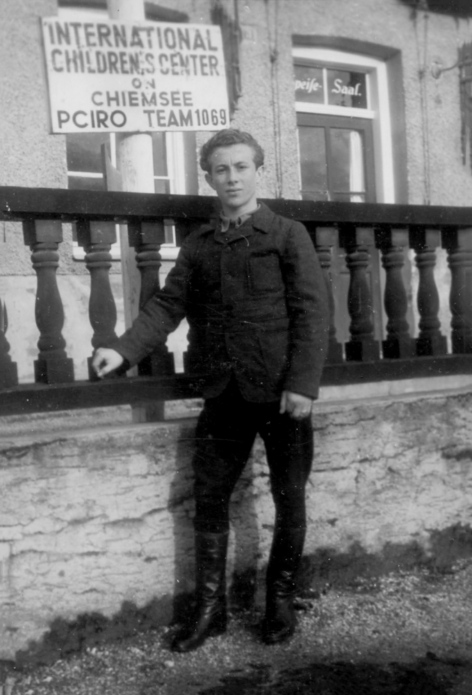 Photo en noir et blanc d'un jeune homme se tenant sur du gazon à l'extérieur et s'appuyant sur une balustrade surélevée en bois qui est derrière lui. Une affiche en arrière-plan inique “INTERNATIONAL CHILDRENS CENTRE”.