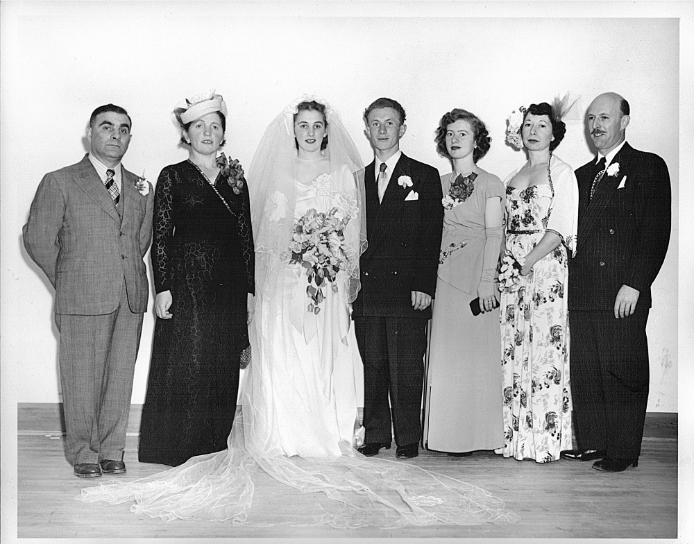 Black-and-white photograph of a group of seven people, standing in a line and smiling at the camera. The couple in the middle appears to be celebrating their wedding day, with the woman dressed in a long white gown, veil, and holding a bouquet of flowers. The men in the group wear suits, and the other three women wear dresses.