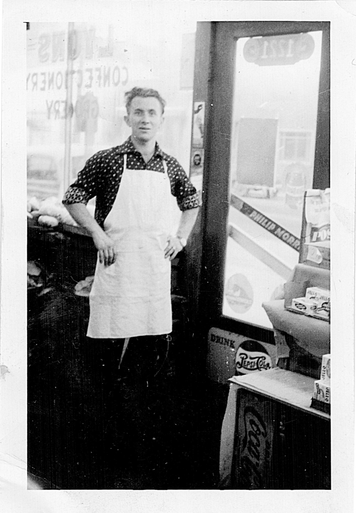 Black-and-white photograph of man standing inside a grocery store. He has his hands on his hips and wears a white apron, standing near the entrance door of the store. The window behind him is printed backwards with the words ‘LYONS CONFECTIONARY STORE’.