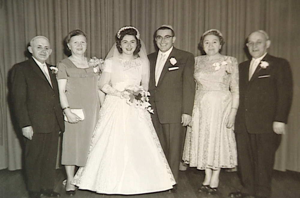 Black-and-white photograph of a group of six people, standing in a line and smiling at the camera. The couple in the middle appears to be celebrating their wedding day, with the woman dressed in a long white gown, veil, and holding a bouquet of flowers. The three men in the group wear suits, and the other two women wear dresses.