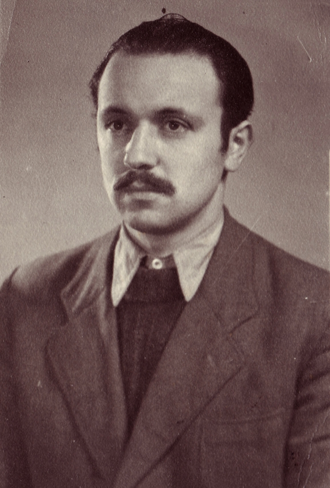 Sepia-toned portrait photograph of a man, pictured from the chest up, looking towards the left of the camera. He wears a suit and tie, and his hair is combed back. He has a moustache.