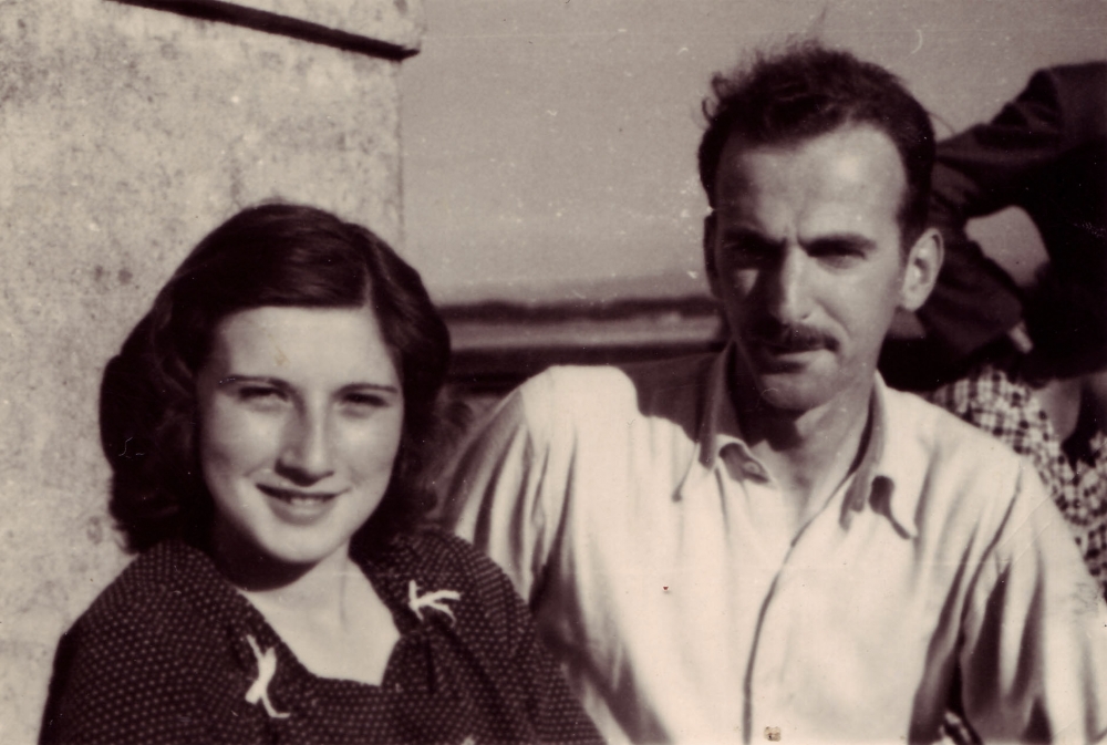 Black-and-white photograph of a man and woman, sitting together outdoors. The woman has shoulder-length brown hair, and the man has a moustache, wearing a light collared shirt.