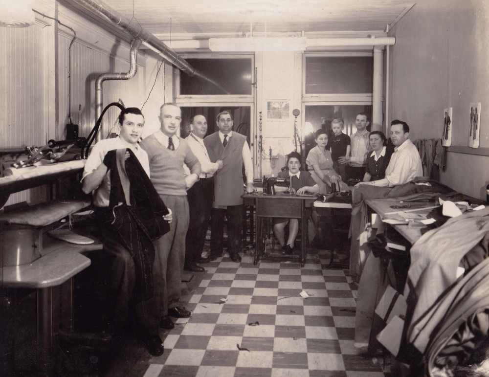 Black-and-white photograph of a group of about ten people standing or sitting together in a room with two windows at the back. The group looks at the camera. Piles of fabric and clothing are on the right, and the floor is checkered.