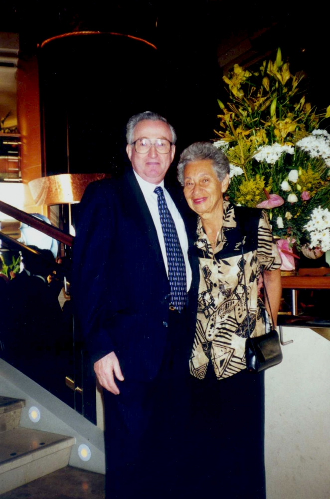 Colour photograph of an elderly couple standing and smiling together, arm-in-arm. The man wears a suit, and the woman wears a printed blouse. They stand in front of a large arrangement of flowers.