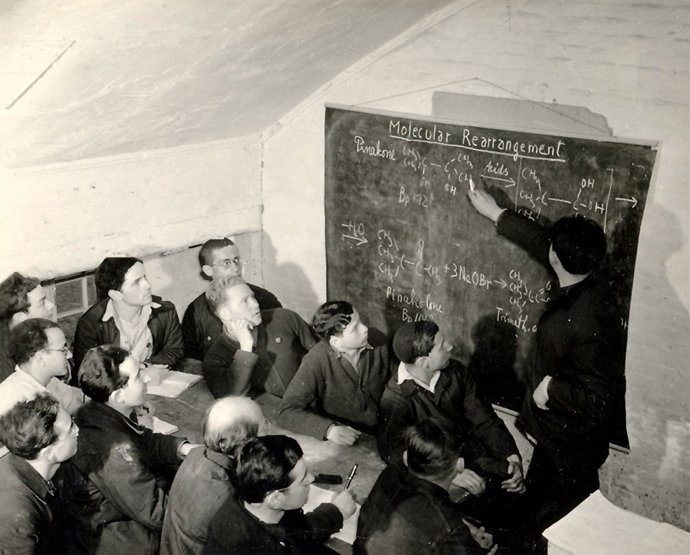 Photo en noir et blanc d'un groupe d'environ douze hommes regardant un tableau. Un des hommes est debout avec un morceau de craie et pointe vers une équation écrite sur le tableau. Les autres hommes sont assis à la table, certains prennent des notes avec papier et crayon.