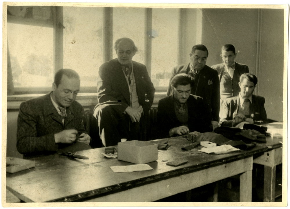 Black-and-white photograph of a group of six men, three standing and three sitting at a table. The men standing are observing the sitting men as they sew fabric with a needle and thread at a table.