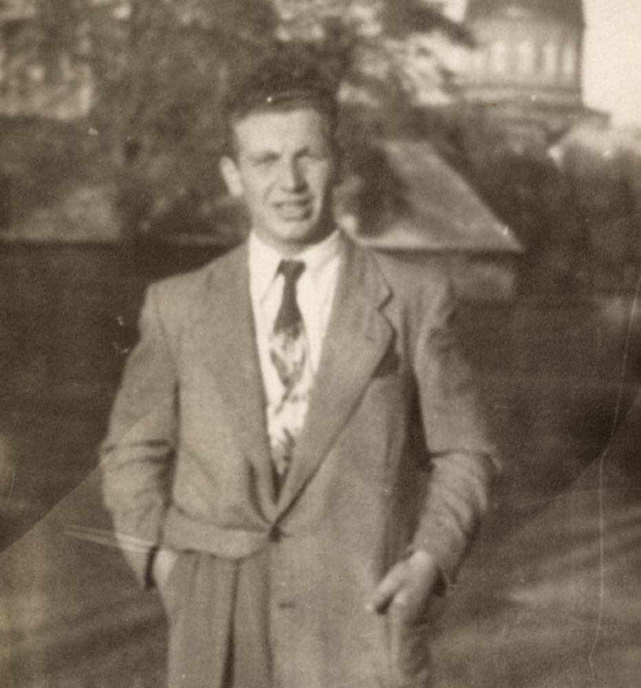 Photo en noir et blanc d'un homme portant un complet et une cravate, debout sur un terrain extérieur. Une tour d'immeuble est visible en arrière-plan.