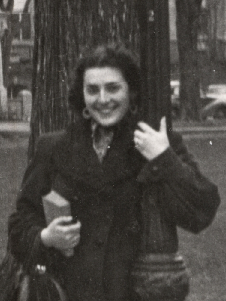 Black-and-white photograph of a woman smiling and leaning against a street light in front of a large field with buildings and cars in the background. She holds a purse and book in her hand.