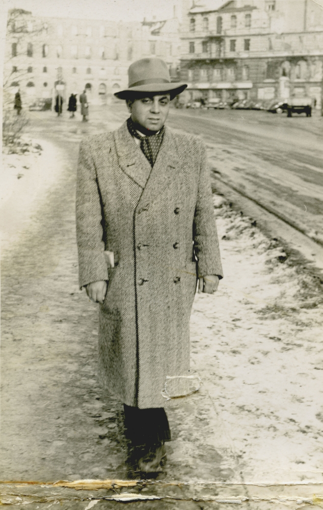 Black-and-white photograph of a man standing outdoors on a snowy sidewalk. The man wears a hat and coat, and there are buildings in the background.