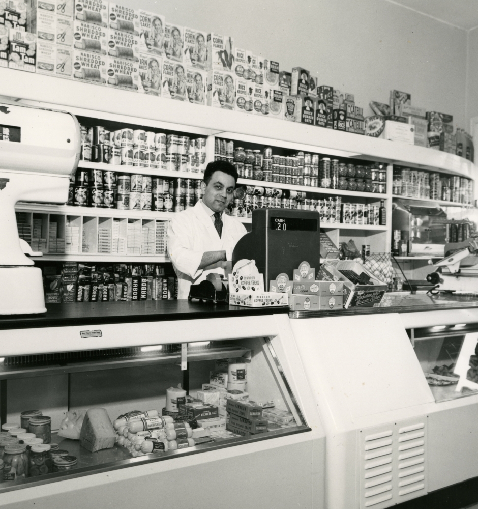 Photo en noir et blanc d'un homme se tenant derrière une caisse enregistreuse dans une grand épicerie. Il se tient devant une étagère remplie de produits et il y a un grand comptoir de présentation devant lui.