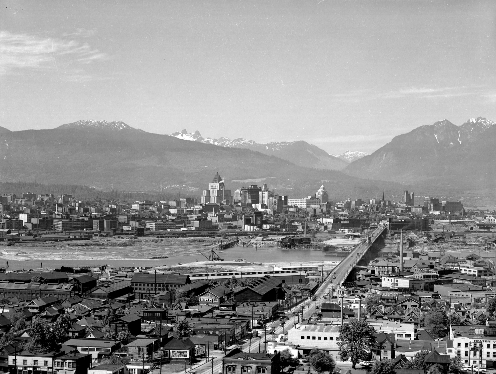 Photo en noir et blanc, vue aérienne d'une ville. Plusieurs bâtiments sont répartis de part et d'autre d'une rivière. Un pont reliant les deux rives se trouve dans la partie droite de la photo. Plusieurs sommets montagneux sont visibles à l'arrière-plan.