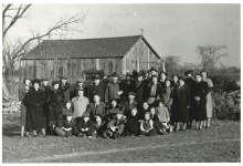 Black-and-white photograph of large group of about 35 men, women, and children, dressed in winter coats and hats, grouped together outdoors on a lawn. There is a barn in the background behind the group.