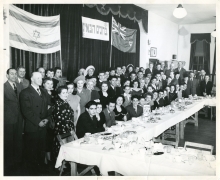 Photo en noir et blanc d'un grand groupe d'environ 70 personnes souriant pour une photo de groupe dans une salle. Le groupe est installé derrière une grande table sur laquelle sont disposés des plats et de la nourriture, et deux drapeaux sont accrochés sur des rideaux en arrière-plan.