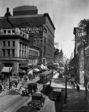 Black-and-white photograph of a busy street scene with vintage cars and streetcars driving up and down a road. Pedestrians walk in either direction on the sidewalks, and there are billboard advertisements posted on buildings’ exterior walls.