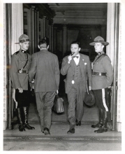 Black-and-white photograph of four men in a large doorway. One man, second from the right, is exiting the room with a briefcase and pipe in his mouth. The man to his left is entering the room. The two men on either side are dressed in RCMP uniforms and look towards the camera.