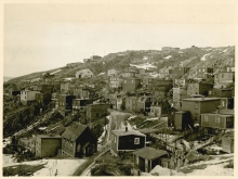 Black-and-white photograph of small residential buildings in a hillside town or village. There is snow on the ground and it appears to be winter.