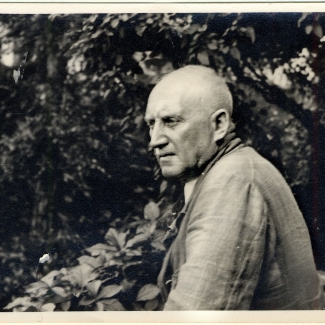 Black-and-white photograph of the side profile of an elderly man standing outdoors, surrounded by leaves and foliage. He looks away in the distance.