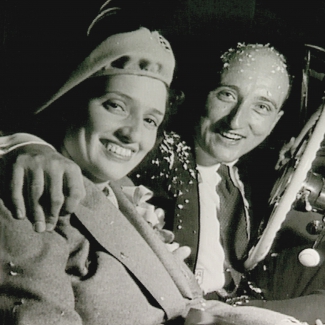 Black-and-white square-shaped photograph of a man and woman sitting together in a car, smiling. The man is covered in confetti and has his arm around the woman.