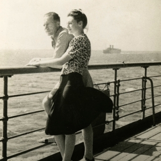 Black-and-white photograph of a woman standing beside a man on the deck of a ship. They both look ahead of them, leaning on the railing of the deck. There is water below them and a ship on the far horizon.