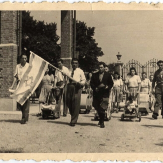 Black-and-white photograph of a parade or march with a large group people walking towards the camera. A man at the front of the crowd carries the flag of Israel, and several women in the background push strollers.