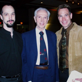 Colour photograph of three men, shown from the waist upwards, smiling at the camera. The elderly man in the middle wears a blue suit and tie, the man on the left wears a black shirt and has facial hair. The man on the right wears a camel jacket and brown shirt with tie.