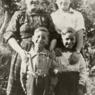 Photo en noir et blanc de deux femmes debout derrière deux jeunes jumeaux, les quatre sourient à la caméra. Ils sont à l'extérieur et il semble que ce soit l'été puisque les manches des femmes sont roulées et que les garçons portent des shorts.